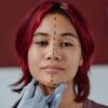 Face of pretty teenage girl with purple hair standing in front of camera while plastic surgeon in protective gloves drawing liftmark preparing for Harmonization Facial Surgery