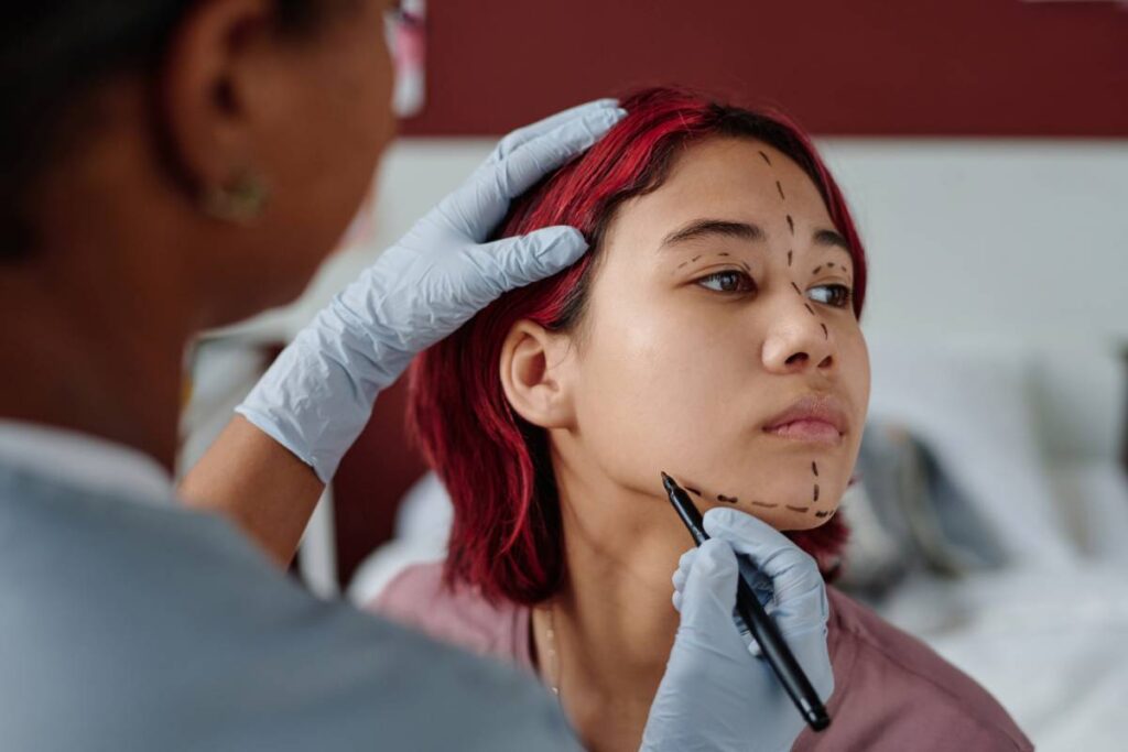 Gloved hands of young professional plastic surgeon with highlighter drawing liftmarks on face of cute teenage girl in clinics