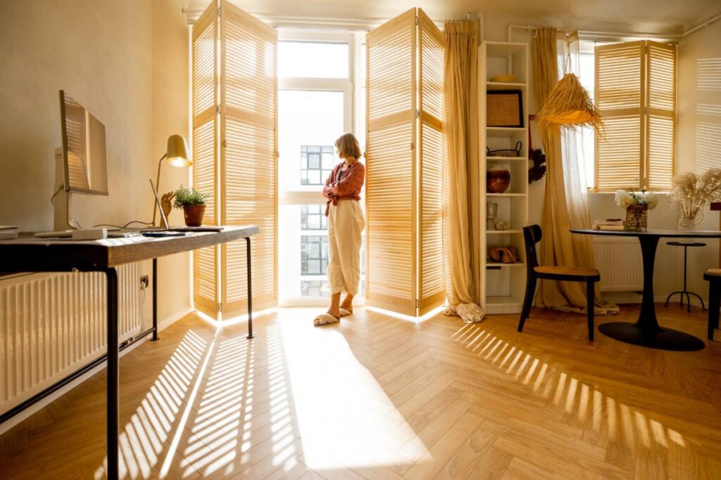 Young woman stands alone by the window blinds at cozy and sunny living room of modern apartment in beige tones. Quarantine, loneliness and life at home