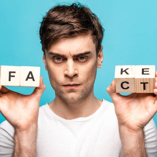 angry man showing wooden cubes with fake fact lettering and looking at camera isolated on blue